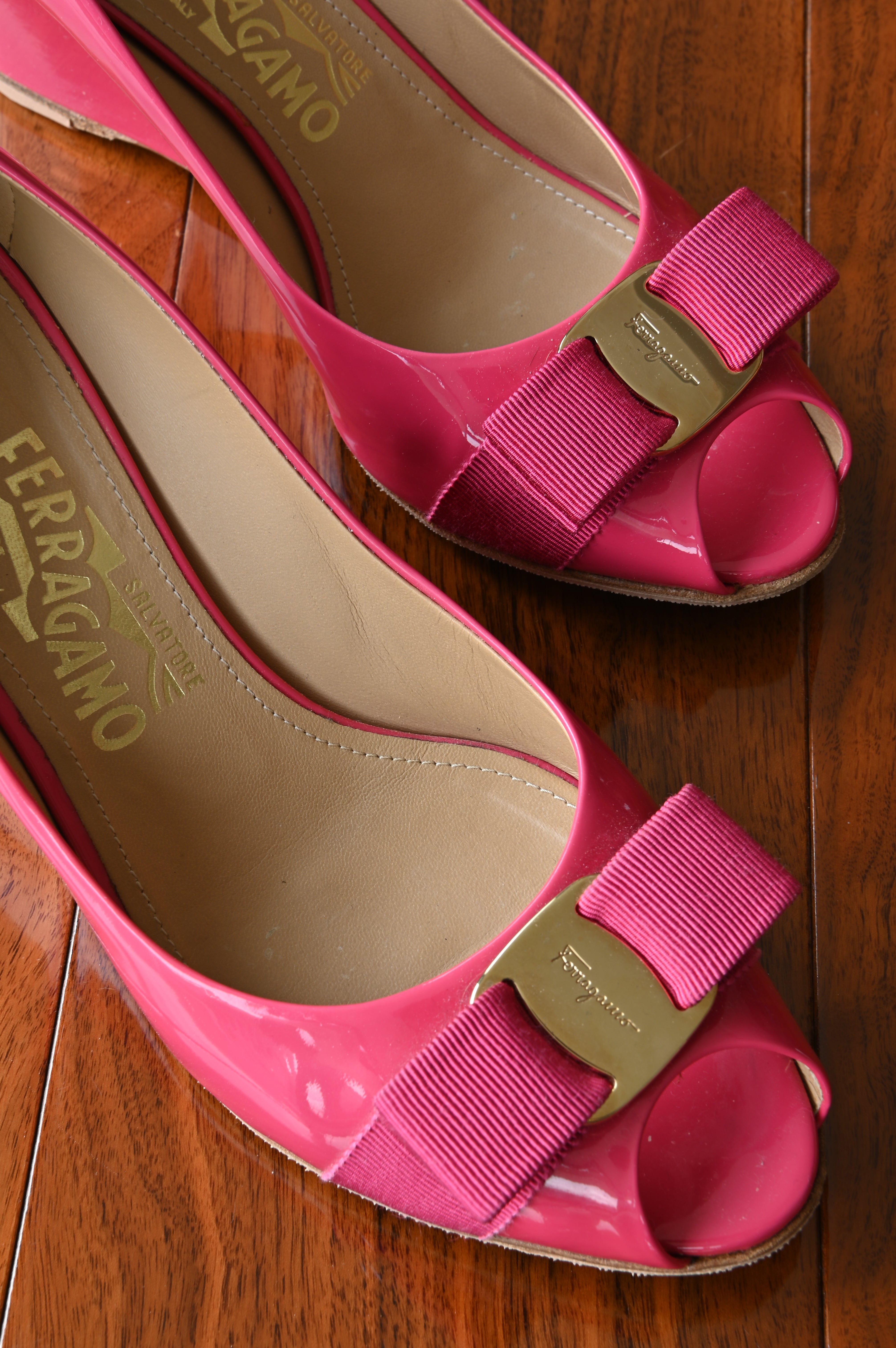 Close-up of the signature Vara grosgrain bow, gold-tone Ferragamo logo plaque, and glossy pink patent leather on the authentic Salvatore Ferragamo wedge pumps.
