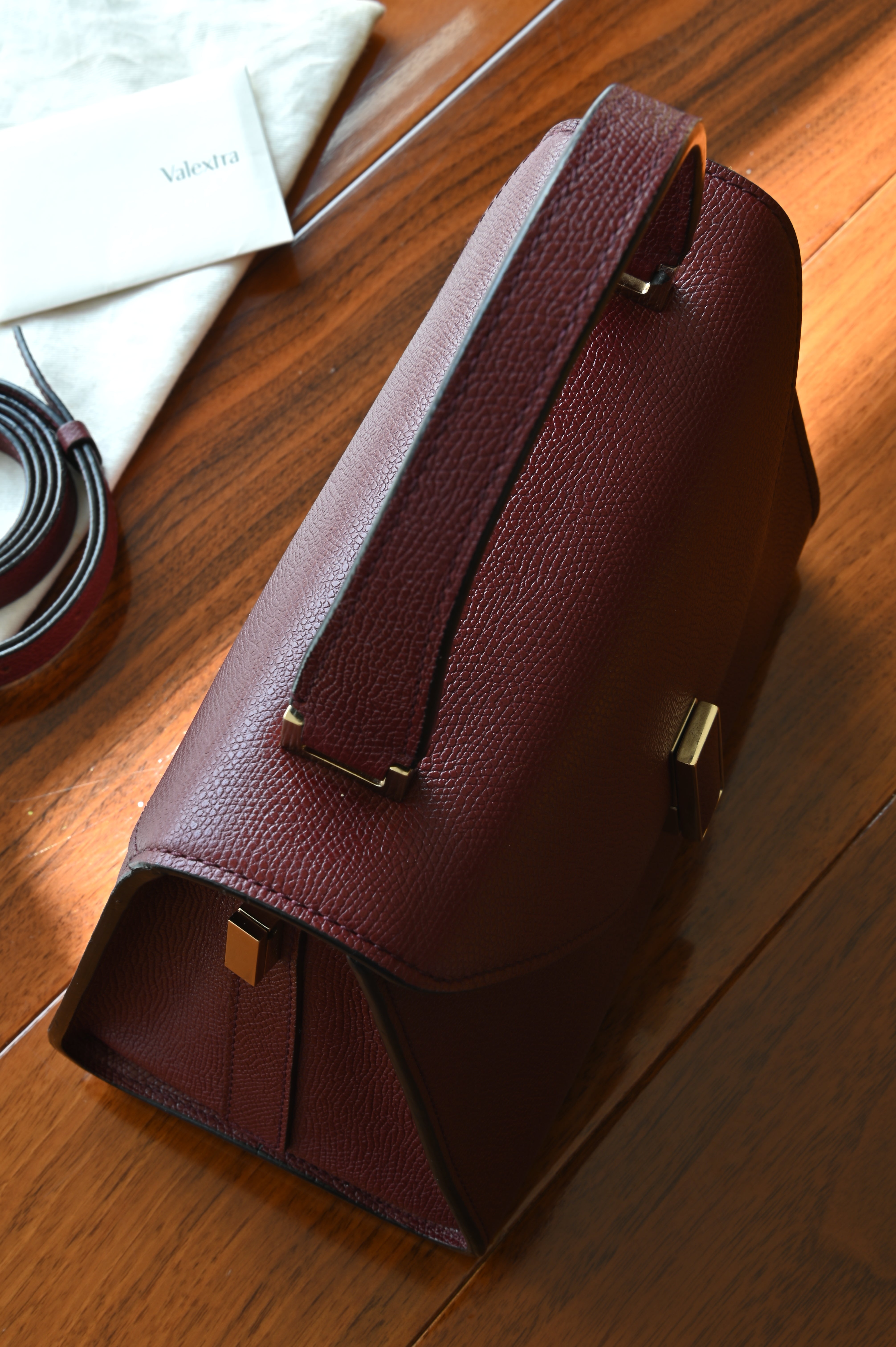 Close-up of the Valextra Iside top handle, detachable shoulder strap, and original dust bag with accompanying authenticity card.